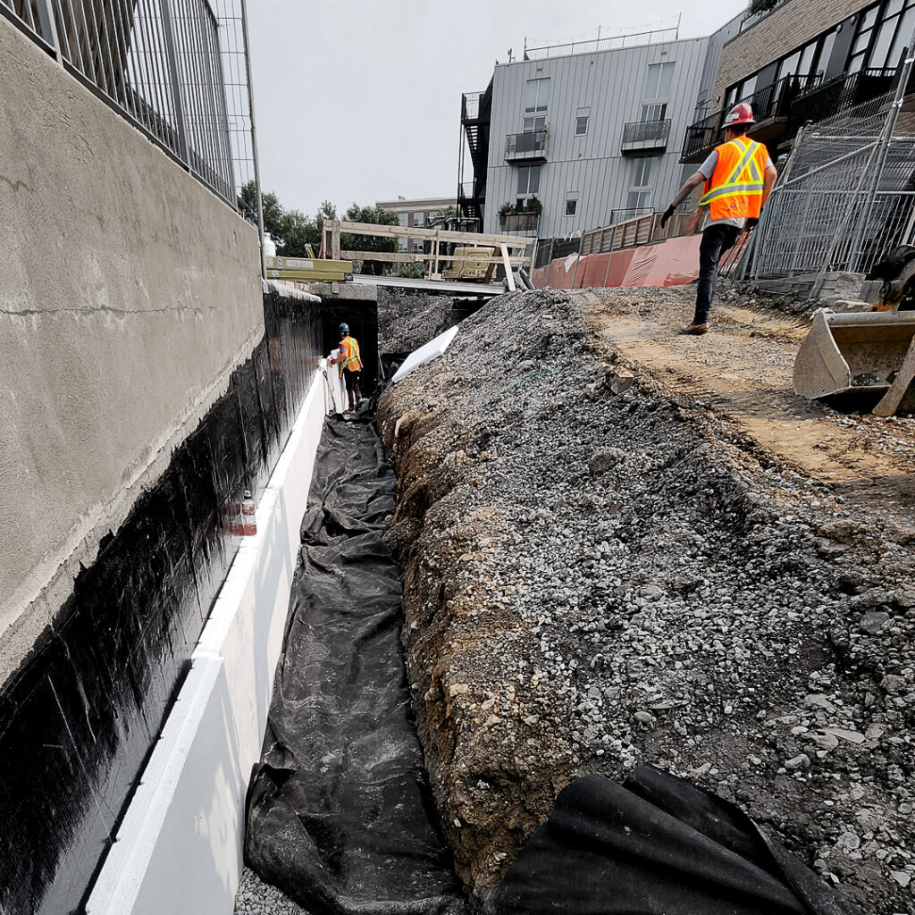 des ouvriers de l'équipe d'excavation Gauthier finissent l'installation d'une membrane isolante autour de la fondation d'une structure de béton.